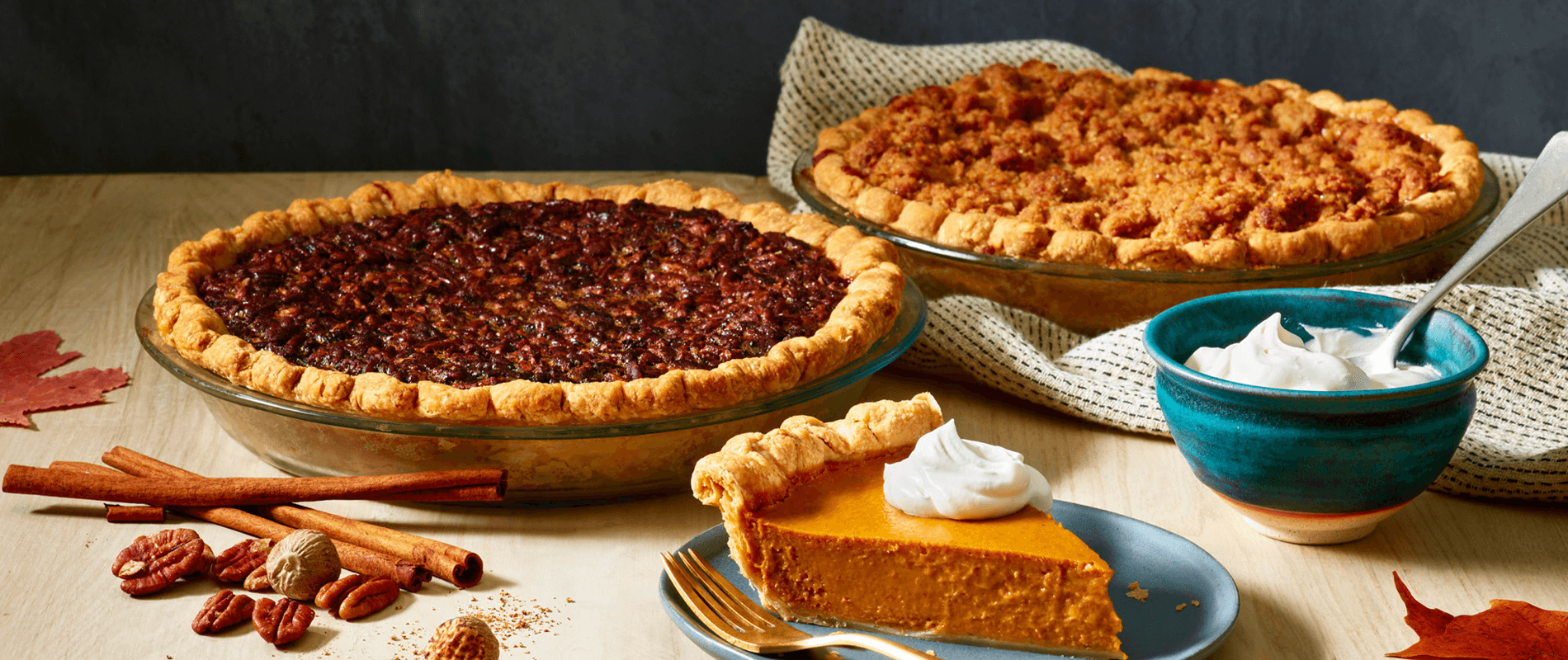 From left to right a whole pecan pie, a slice of pumpkin pie with whipped cream, a whole apple pie and a bowl of freshly whipped cream, surrounded by cinnamon sticks, pecans, powdered spices and maple leaves 