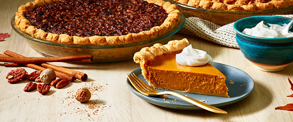 From left to right a whole pecan pie, a slice of pumpkin pie with whipped cream, a whole apple pie and a bowl of freshly whipped cream, surrounded by cinnamon sticks, pecans, powdered spices and maple leaves 